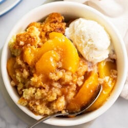 Peach Cobbler in a white bowl with a spoon and a scoop of vanilla ice cream on top.