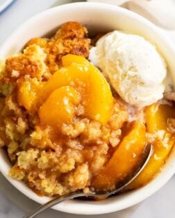 Peach Cobbler in a white bowl with a spoon and a scoop of vanilla ice cream on top.