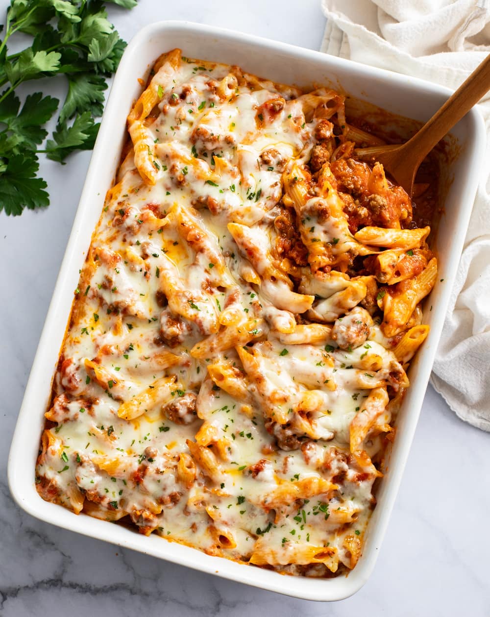A white baking dish filled with Ground Beef Casserole with melted mozzarella on top and a spoon on the side.