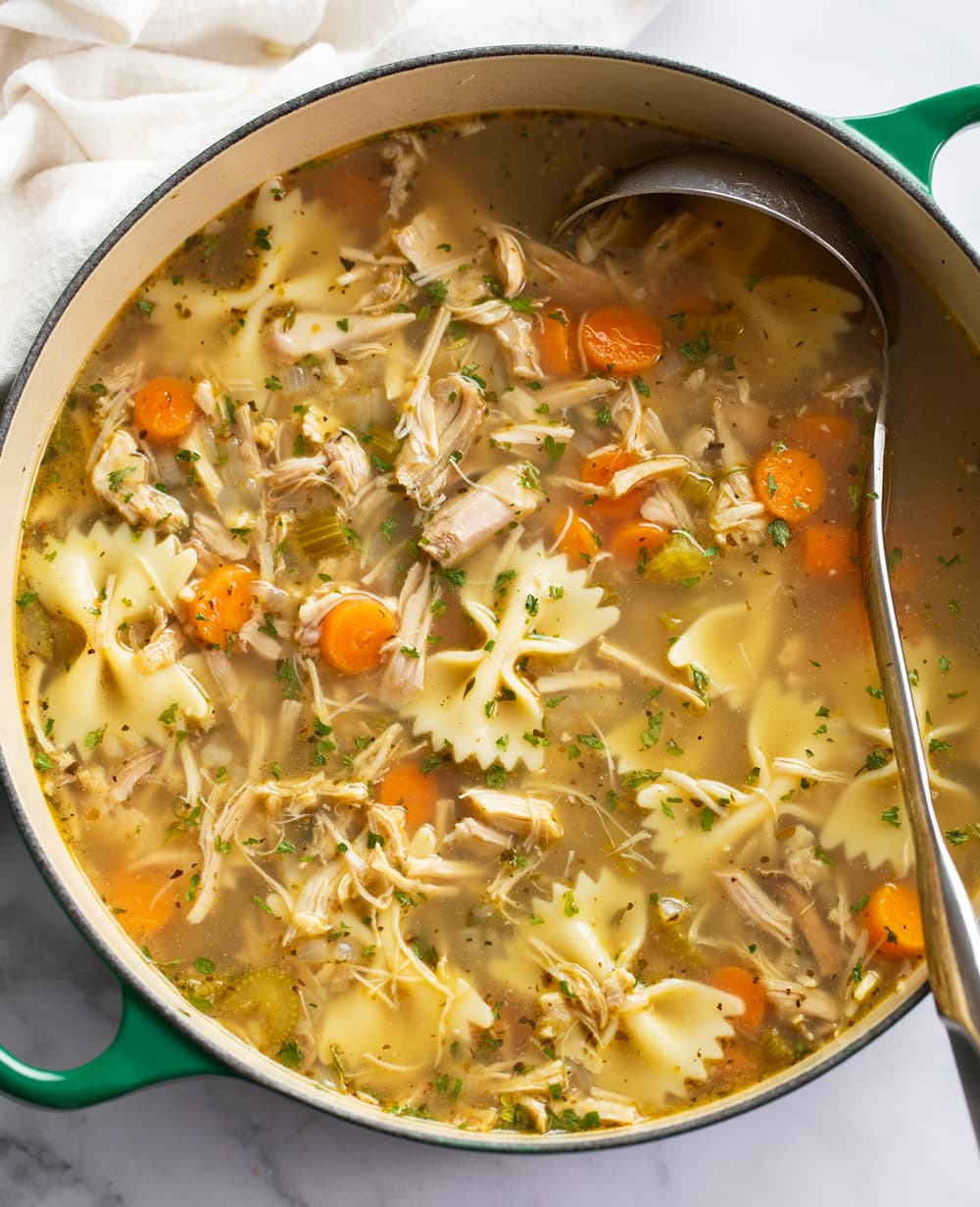 A soup pot filled with Turkey Soup with vegetables, bowtie pasta, and a ladle on the side.