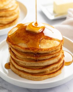 A stack of Homemade Pancakes on a white plate with syrup being poured on top.