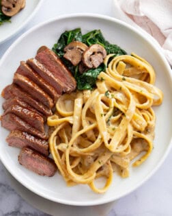 Fettuccine in a creamy Alfredo sauce on a white plate next to Steak, spinach, and mushrooms.