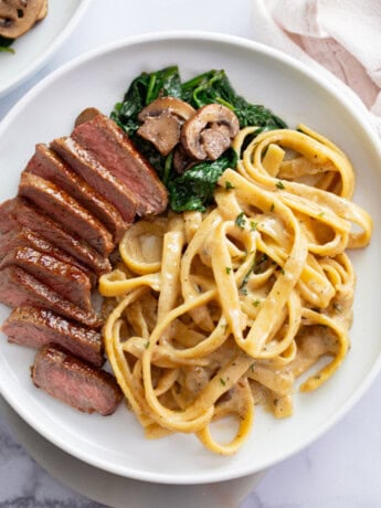 Fettuccine in a creamy Alfredo sauce on a white plate next to Steak, spinach, and mushrooms.