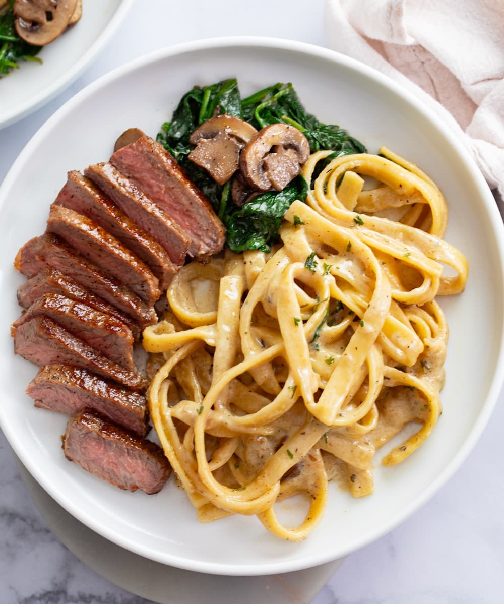 Fettuccine in a creamy Alfredo sauce on a white plate next to Steak, spinach, and mushrooms.