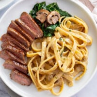 Steak Pasta on a white plate with a creamy alfredo sauce, spinach, and mushrooms.