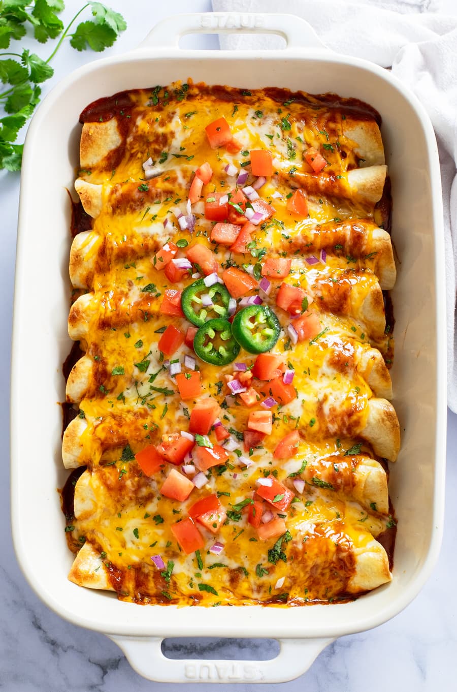 Cheesy Chicken Enchiladas in a casserole dish with diced tomatoes, cilantro, onions, and jalapenos on top.