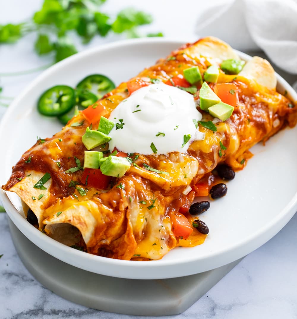 Chicken Enchiladas on a white plate with sour cream and diced avocados on top.