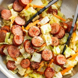 Cabbage and Sausage in a skillet with carrots and kitchen tongs.