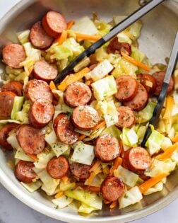 Cabbage and Sausage in a skillet with carrots and kitchen tongs.