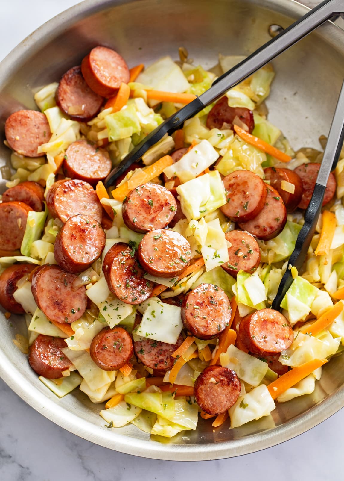 Cabbage and Sausage in a skillet with carrots and kitchen tongs.
