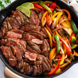 A skillet filled with Steak Fajitas with slices of lime on the side.