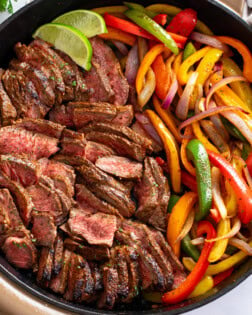 A skillet filled with Steak Fajitas with slices of lime on the side.