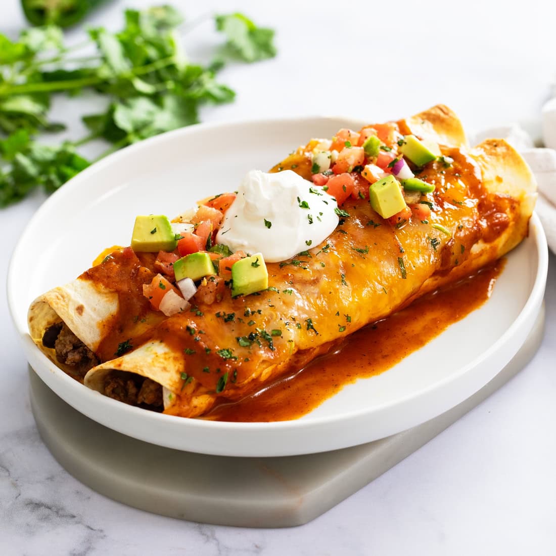 Two beef enchiladas on a white plate with a dollop of sour cream on top with diced avocados and tomatoes.