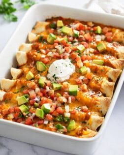 Beef Enchiladas in a white baking dish topped with cheese, tomatoes, avocados, and sour cream.