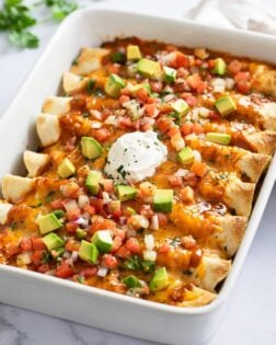 A white casserole dish filled with beef enchiladas with tomatoes, avocados, and sour cream on top.