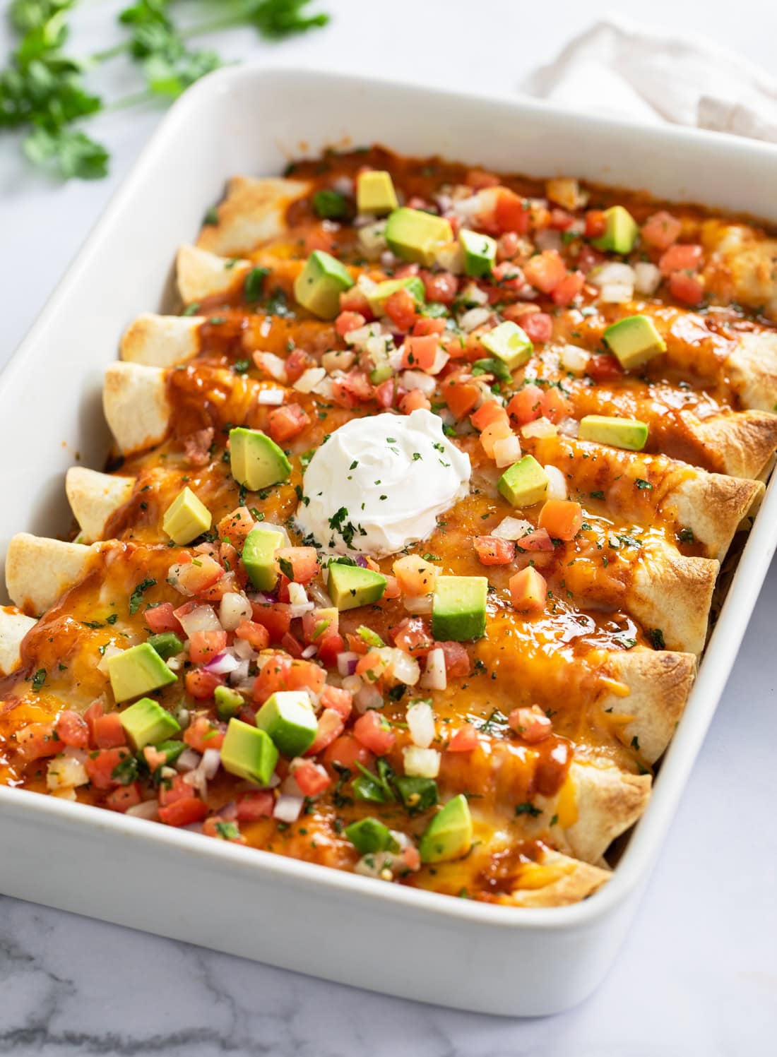 A white casserole dish filled with beef enchiladas with tomatoes, avocados, and sour cream on top.