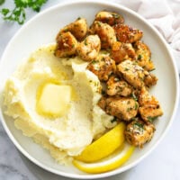 Chicken Bites on a next to mashed potatoes on a white plate with lemon wedges on the side.