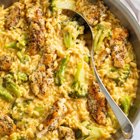 Chicken and Orzo in a skillet with a creamy cheese sauce and broccoli with a spoon on the side.