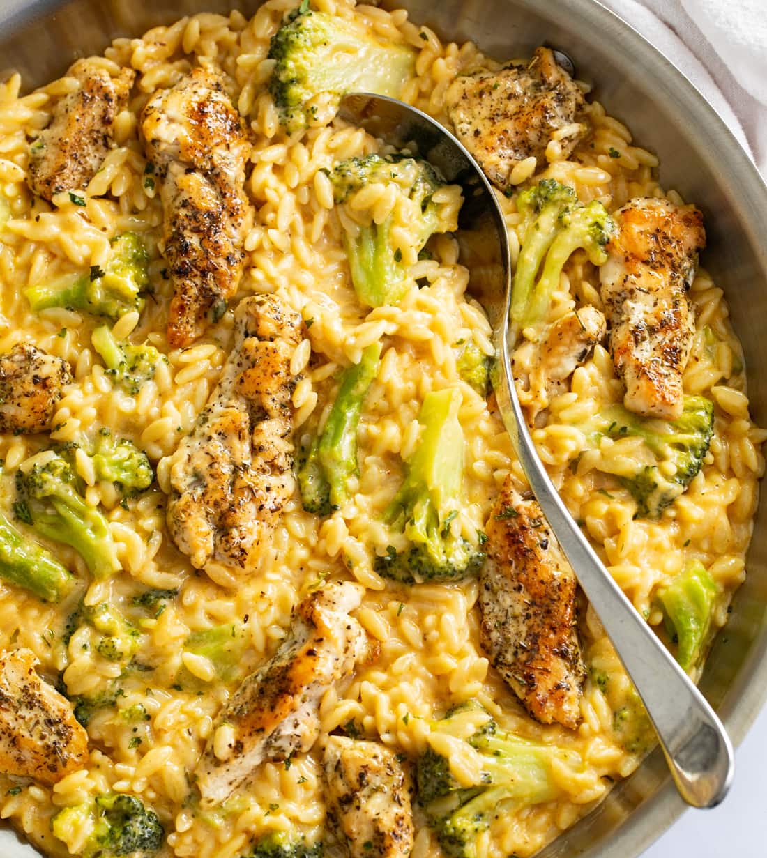 Chicken and Orzo in a skillet with a creamy cheese sauce and broccoli with a spoon on the side.