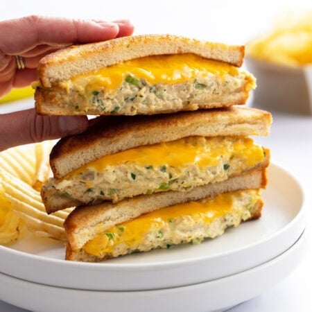 A hand taking the top Tuna Melt from a stack of 3 on a white plate with melted cheese.