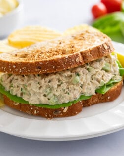 A Tuna Salad Sandwich with toasted bread on a white plate with a pickle and chips in the background.