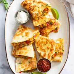A white tray with sliced Chicken Quesadillas on top with sour cream, salsa, and lime wedges.