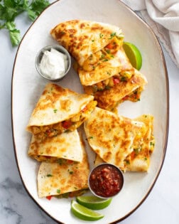 A white tray with sliced Chicken Quesadillas on top with sour cream, salsa, and lime wedges.