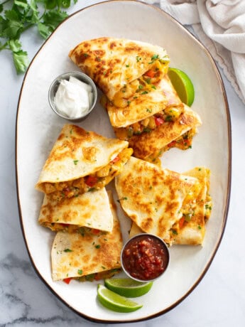 A white tray with sliced Chicken Quesadillas on top with sour cream, salsa, and lime wedges.