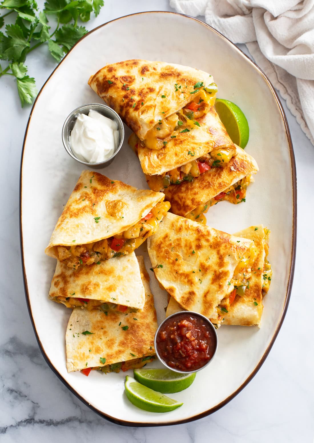 A white tray with sliced Chicken Quesadillas on top with sour cream, salsa, and lime wedges.