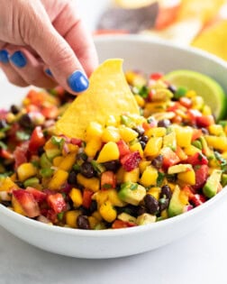 A hand dipping a tortilla chip into a white bowl of Mango Salsa.