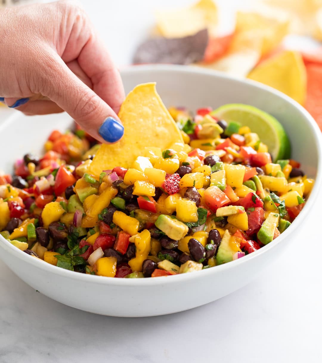 A white bowl of Mango Salsa with a tortilla chip being dipped into it.