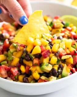 A hand dipping a tortilla chip into a white bowl of Mango Salsa.