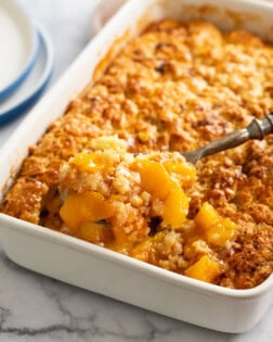 A spoon scooping up Peach Cobbler from a white casserole dish.