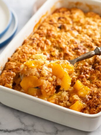 A spoon scooping up Peach Cobbler from a white casserole dish.