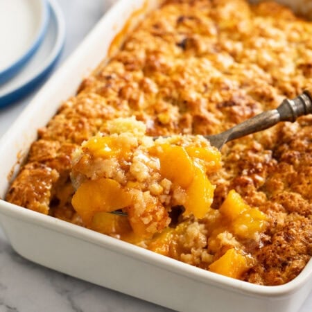 A spoon scooping up Peach Cobbler from a white casserole dish.