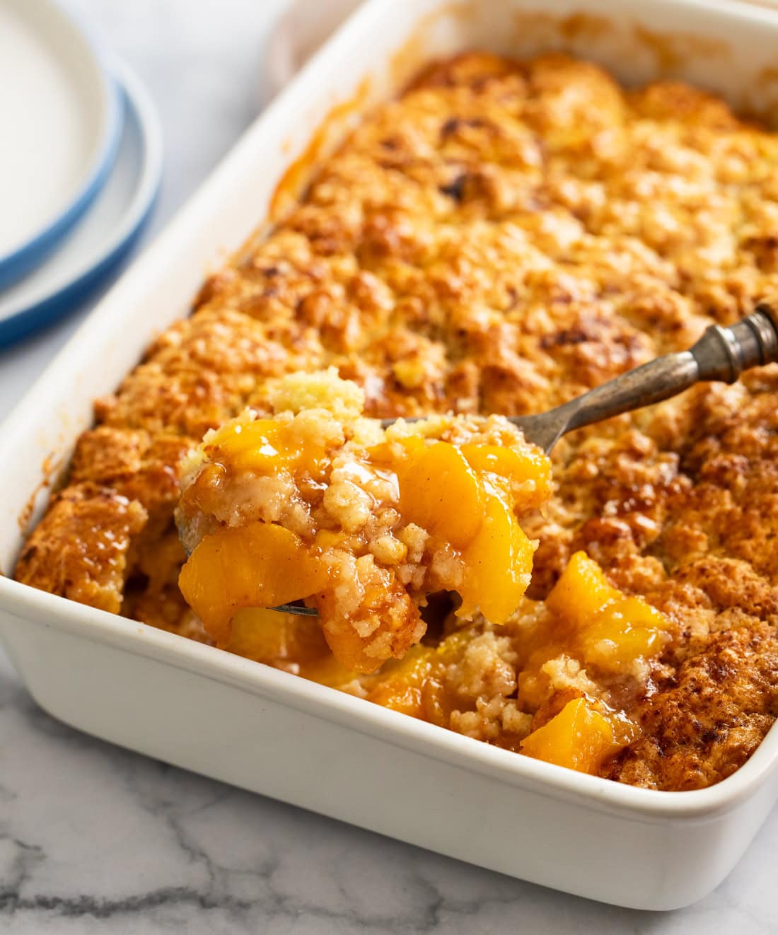 A spoon scooping up Peach Cobbler from a white casserole dish.