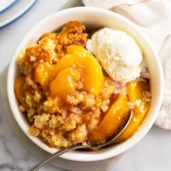 A white bowl of peach cobbler with a fluffy biscuit topping and a scoop of ice cream on the side.