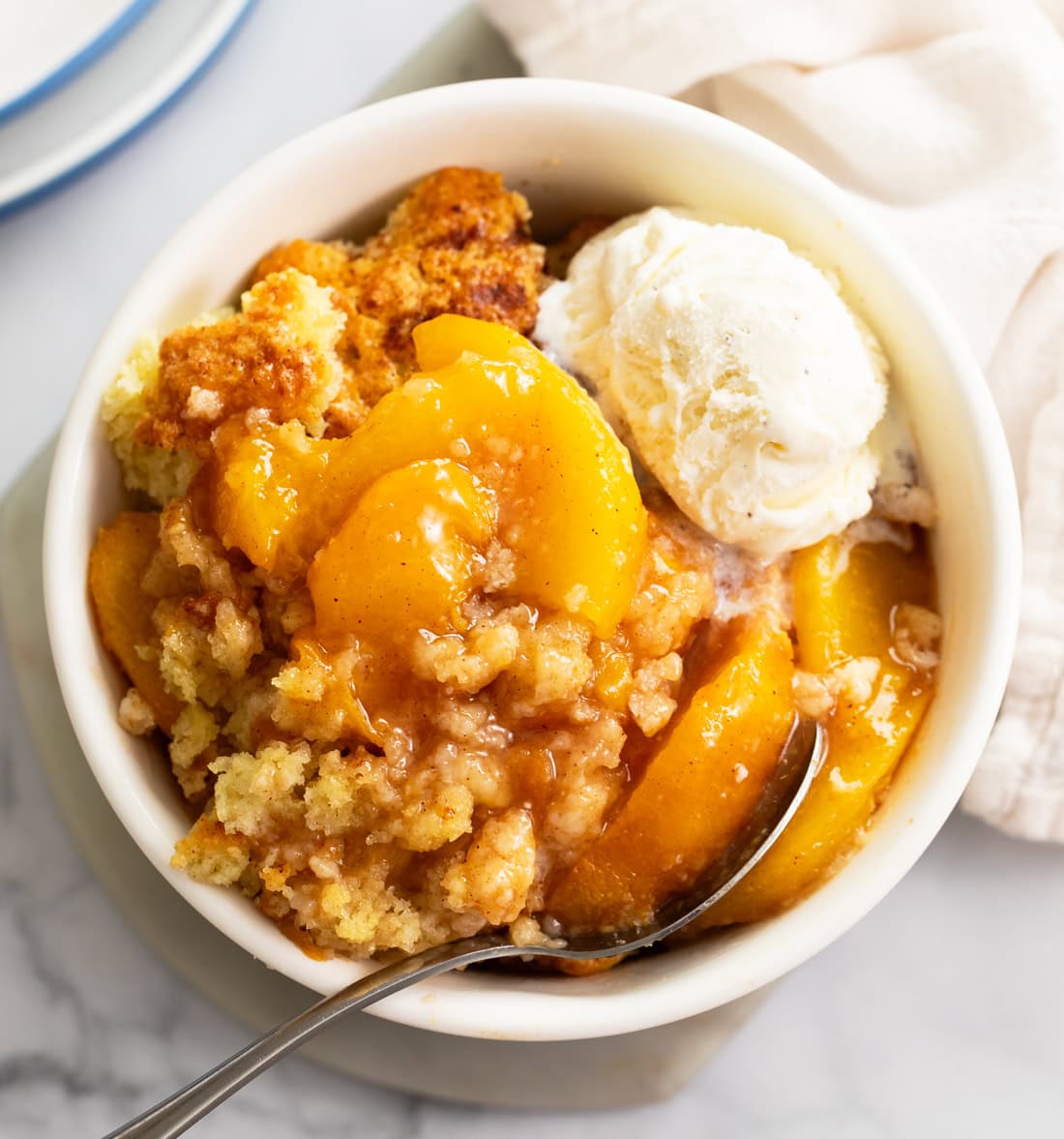 A white bowl of peach cobbler with a fluffy biscuit topping and a scoop of ice cream on the side.