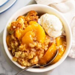 Peach Cobbler in a white bowl with a spoon and a scoop of vanilla ice cream on the side.