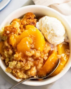 Peach Cobbler in a white bowl with a spoon and a scoop of vanilla ice cream on the side.