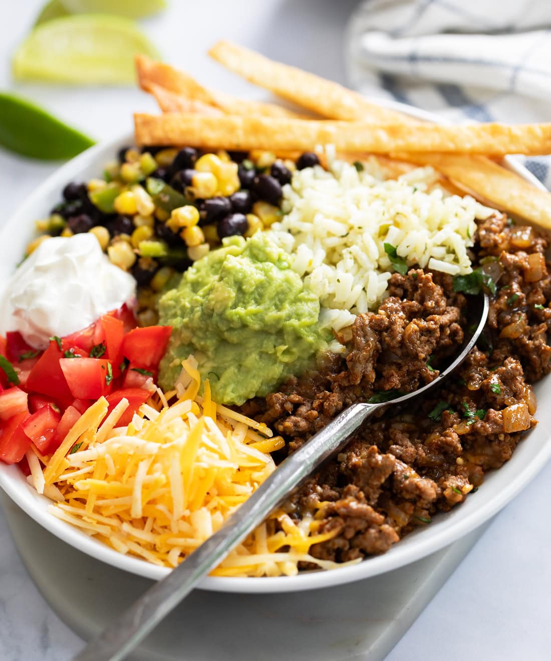 A spoon scooping up seasoned ground beef from a burrito bowl with rice, cheese, beans, and more.