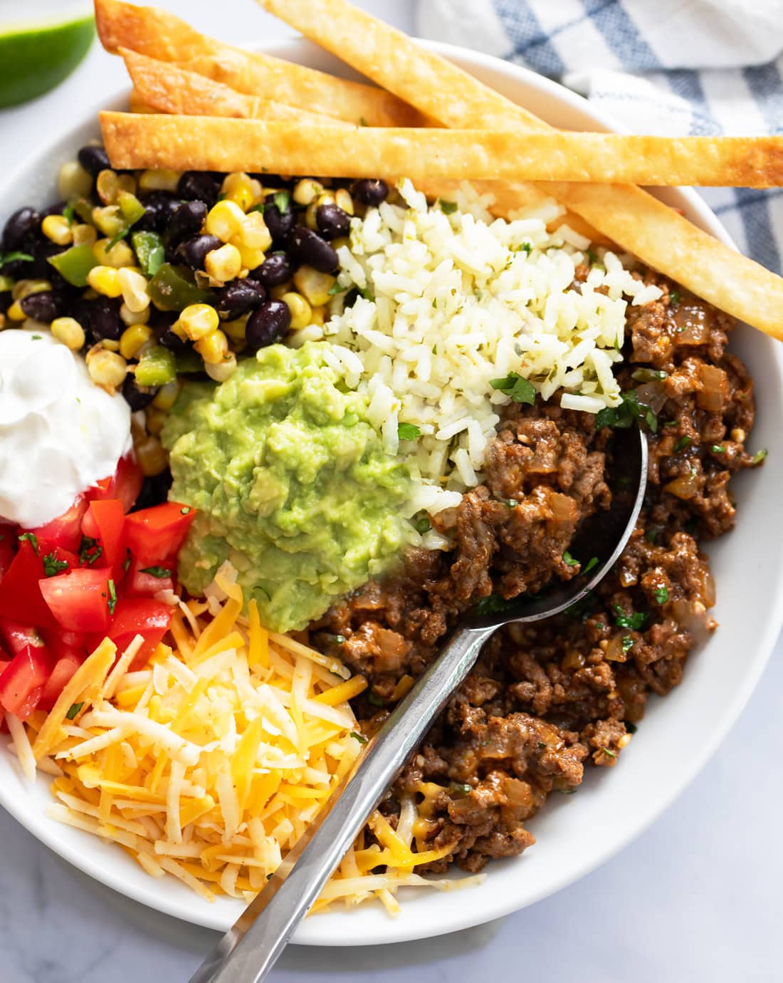 Close up view of the ingredients of a Burrito Bowl, including ground beef, guacamole, rice, beans, corn, tomatoes, cheese, and sour cream.