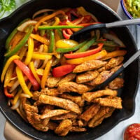 Chicken Fajitas in a cast iron skillet with bell peppers, seasoned chicken, and kitchen tongs.