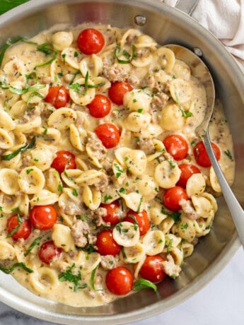 Tomato Basil Pasta in a skillet with fresh tomatoes and basil in a cream sauce with a spoon on the side.