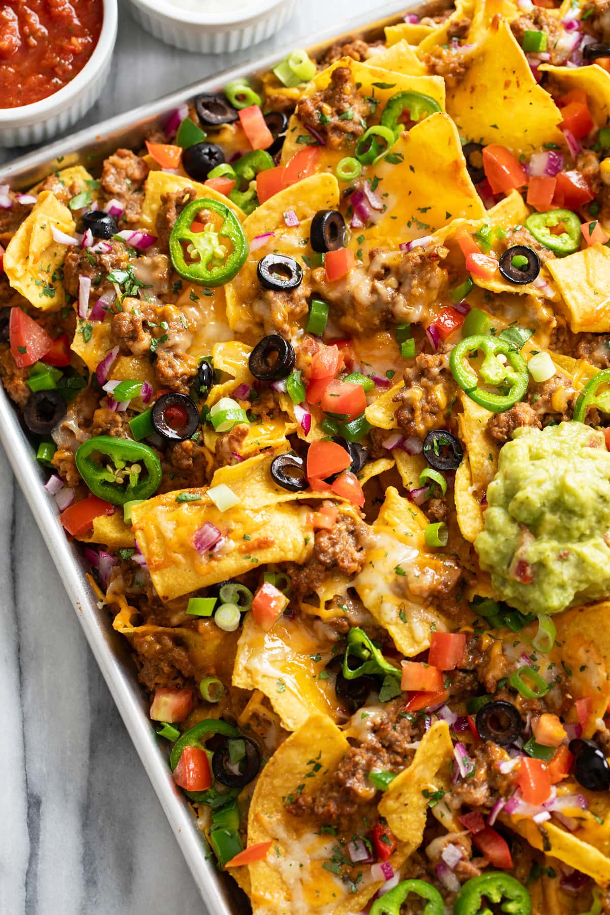 A tray of loaded Nachos on a sheet pan with beef, refried beans, cheese, and a scoop of guacamole on top.