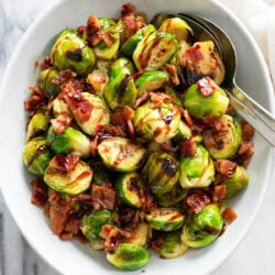 Bacon Brussels Sprouts in a white bowl with a spoon on the side and chopped bacon on top with balsamic glaze.