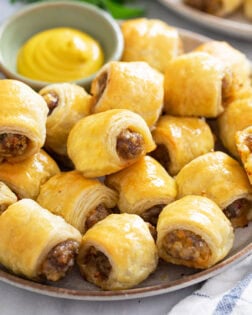 A plate of Sausage Rolls stacked up with mustard in the back for dipping.