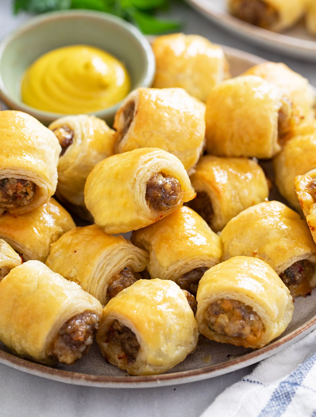 A plate of Sausage Rolls stacked up with mustard in the back for dipping.