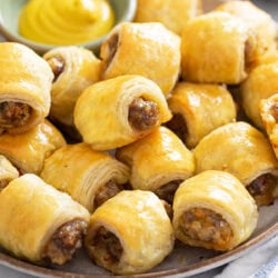 A plate of puff pastry Sausage Rolls with a side of mustard in the background for dipping.