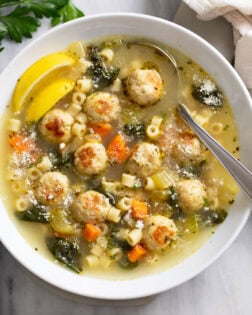 Chicken Meatball Soup in a white serving bowl with lemon wedges and a spoon on the side.